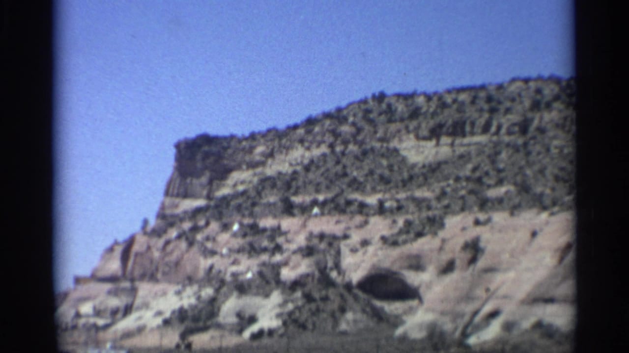 Wide canyon view with layered rock, blue sky, and a cave opening near the road.