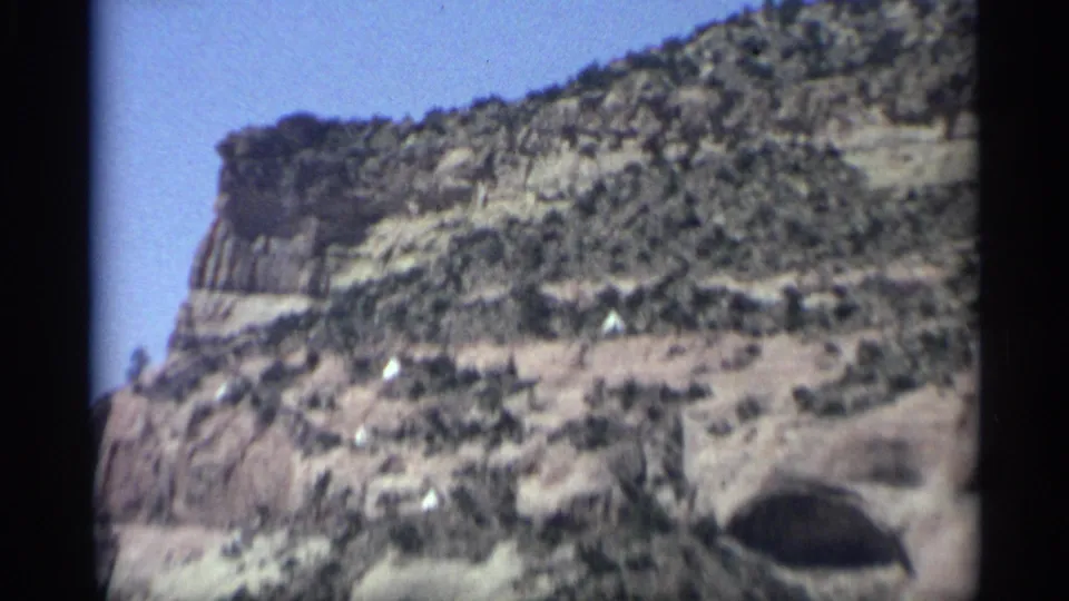 1959 Arizona 8mm frame looking across a rugged canyon slope with scrub vegetation and a cave opening below.