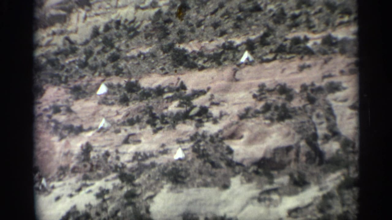 Desert hillside with scattered brush, rock formations, and small white tents.