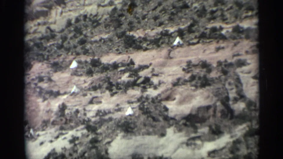 Arizona desert hillside with several small white tents set among brush below a layered rock wall.