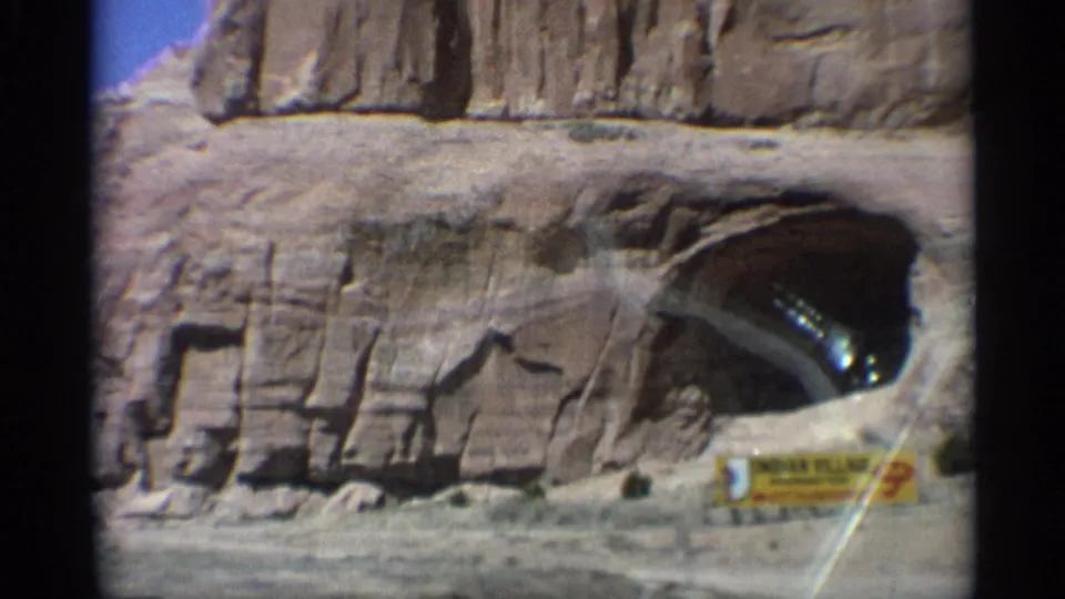 Arizona desert roadside view with a dark sandstone arch or cave entrance, layered cliff face, and a small sign near the lower right.