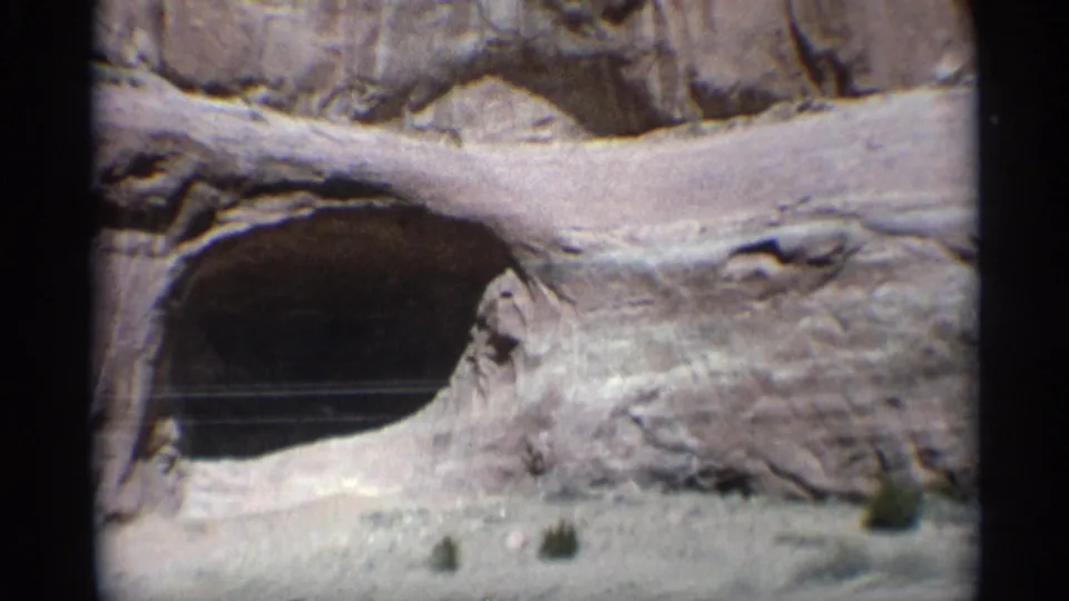 1959 Arizona desert frame centered on a large dark rock arch or cave opening, with roadside pavement and sparse shrubs below.