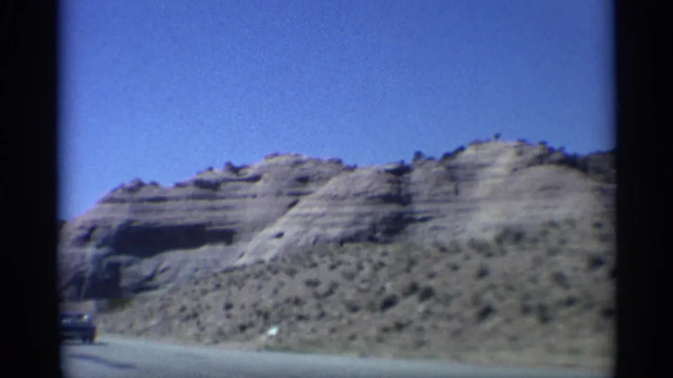 Wide 1959 Arizona home-movie view of a road below pale sandstone hills, with a partial vintage car at the lower left.