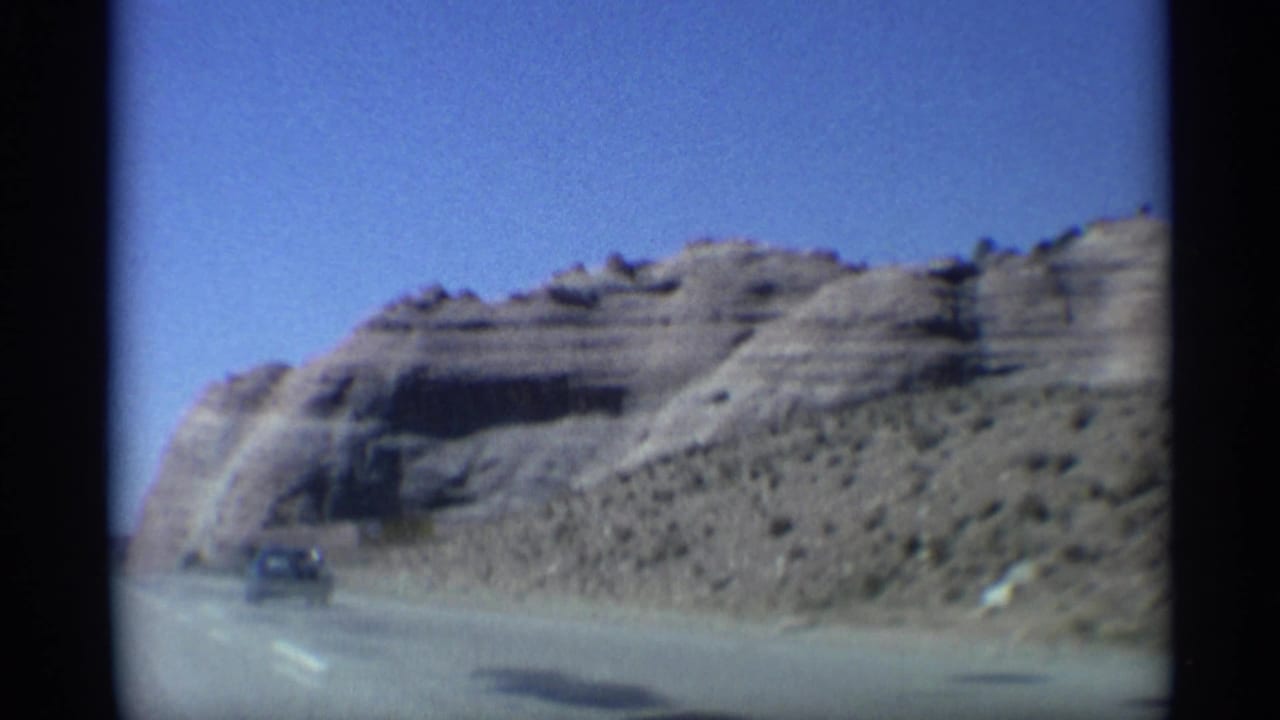 Desert highway beside a rounded cliff with a vehicle approaching in the distance.