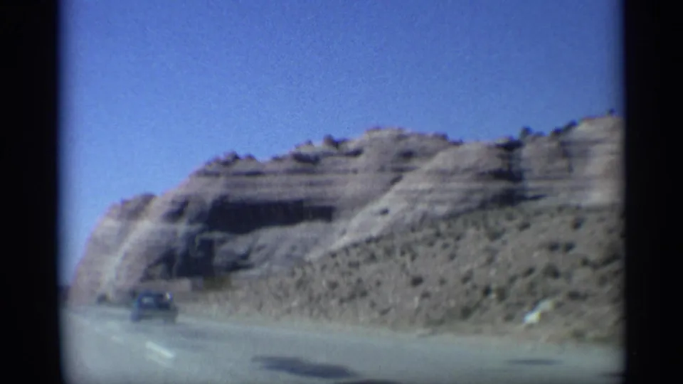 1959 Arizona 8mm road-trip frame showing a desert highway, pale cliff face, sparse scrub, and a distant vehicle under blue sky.