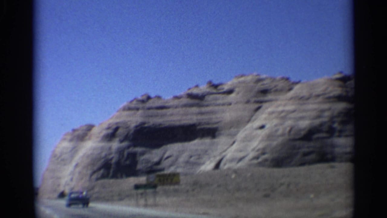 1959 Arizona travel footage frame with a blue truck, roadside Indian City sign, desert shoulder, and cliff face in the background.
