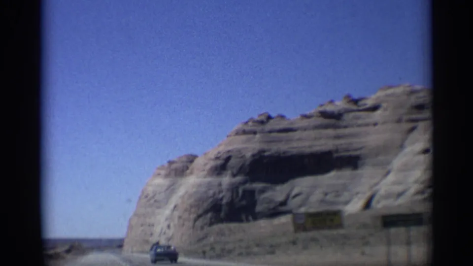 Arizona 8mm frame from 1959 showing a truck on the road, a rounded cliff, and partially readable roadside signs from a moving vehicle.
