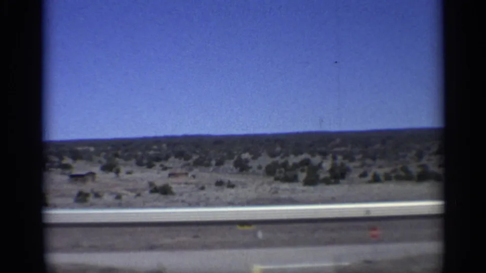 1959 Arizona road-trip frame showing highway guardrail, flat desert scrub, distant horizon, and clear blue sky from a moving-car viewpoint.
