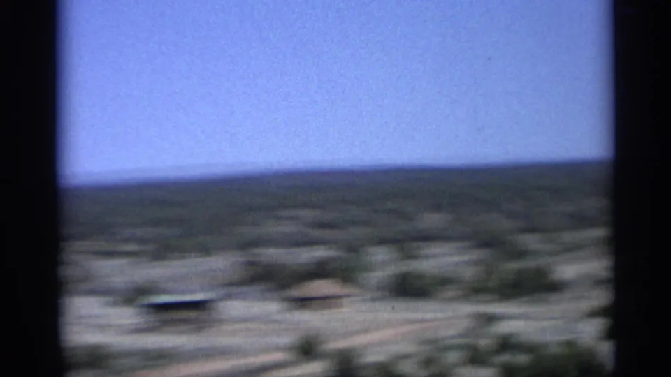 Motion-blurred 1959 Arizona road-trip frame showing low desert buildings, scrub vegetation, and open horizon.