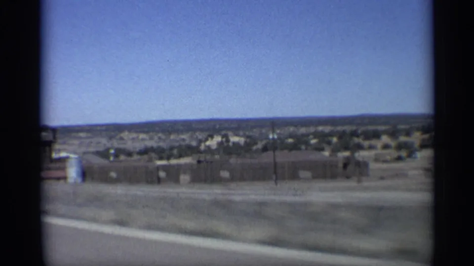 1959 Arizona road view with fencing, low structures, sparse desert vegetation, distant hills, and open sky.