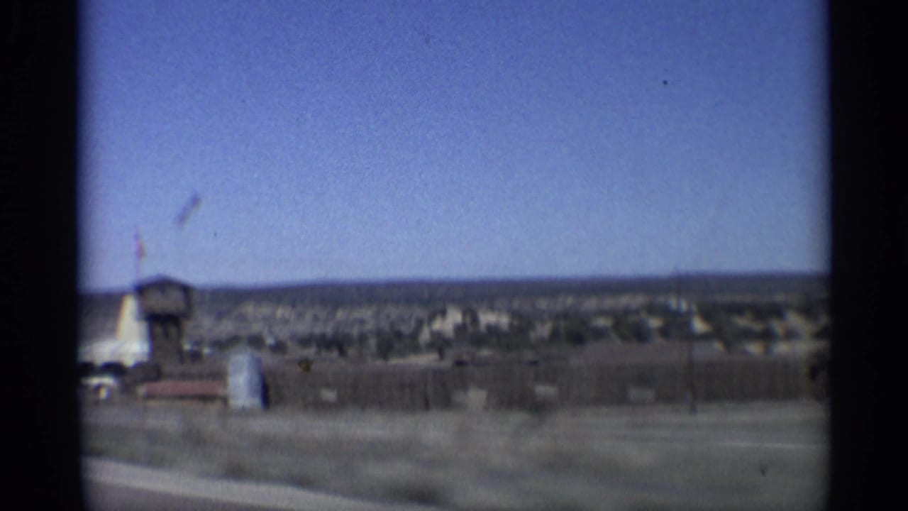 Moving highway view of a desert roadside compound with a tower, flag, low buildings, and open sky.