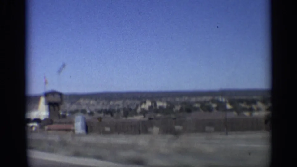 1959 Arizona 8mm frame showing a drive-by view of a remote desert roadside compound with a small tower, flag, and low structures.