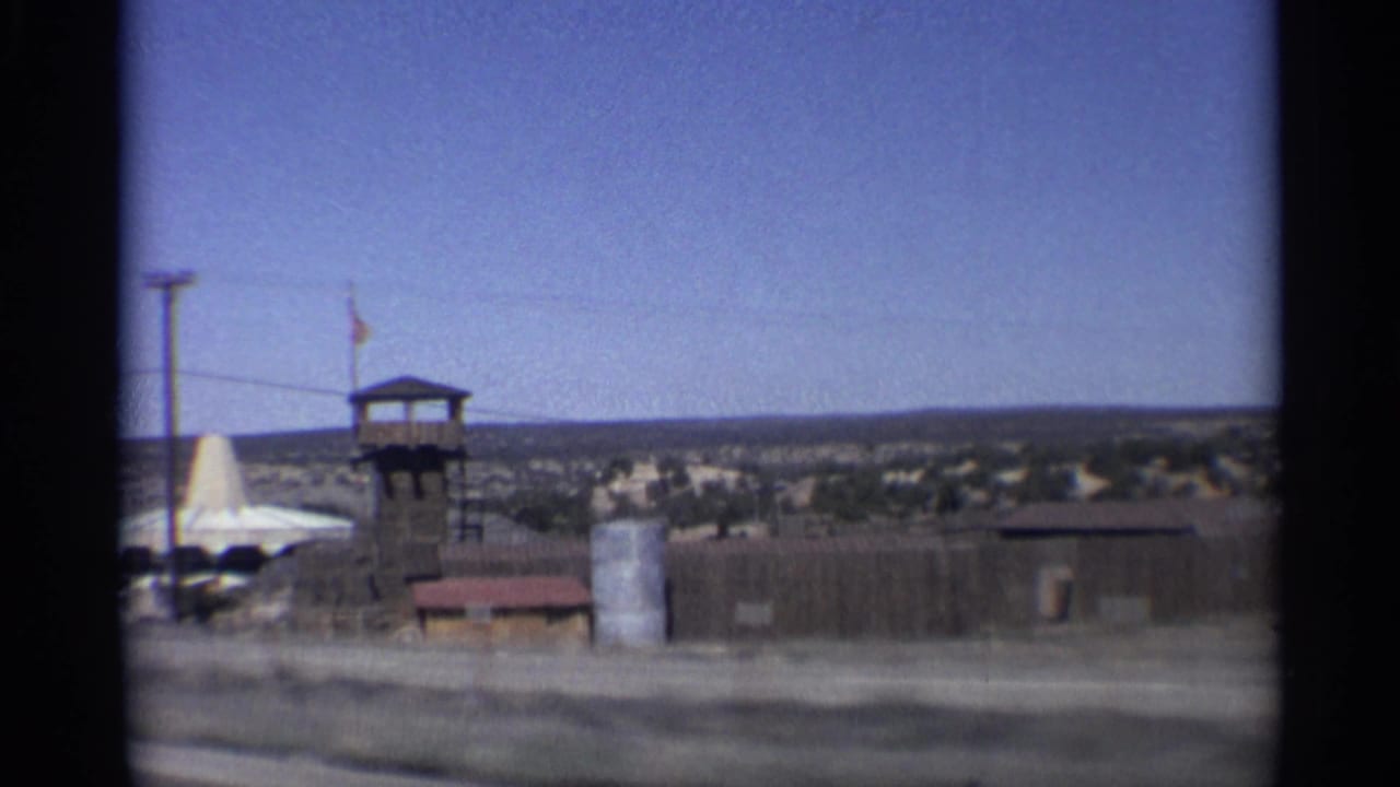 Roadside tower with flag, low buildings, fencing, and desert terrain seen from a moving vehicle.