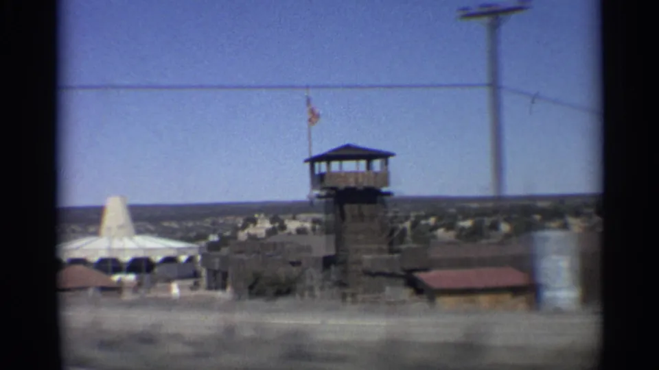 1959 Arizona roadside frame showing a tower with flag, conical structure, utility pole, and low buildings blurred by vehicle motion.