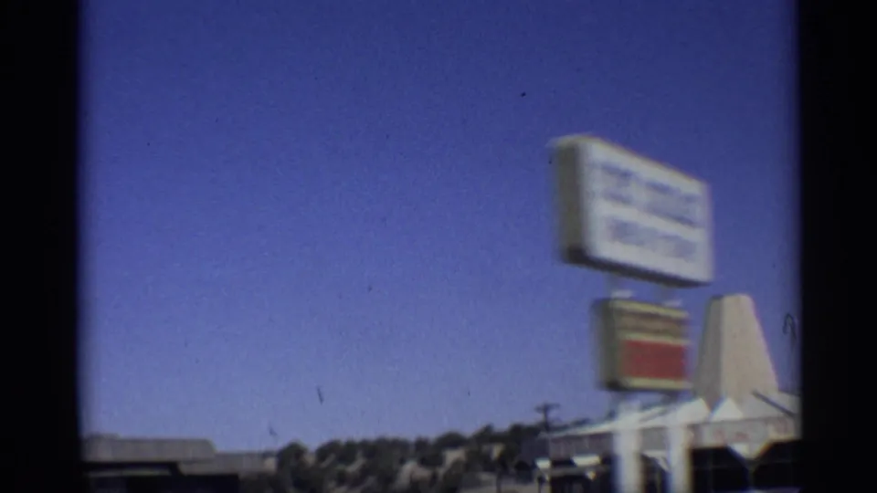 1959 Arizona drive-by frame with an unreadable roadside sign, a conical structure, low building edges, and open desert sky.