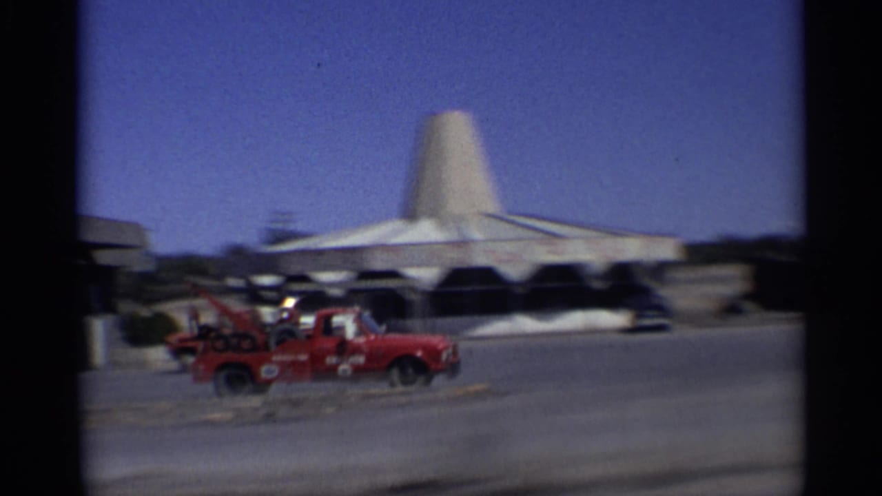 1959 Arizona home-movie frame showing a red tow truck, parked cars, and a distinctive conical-roof building beside a road.