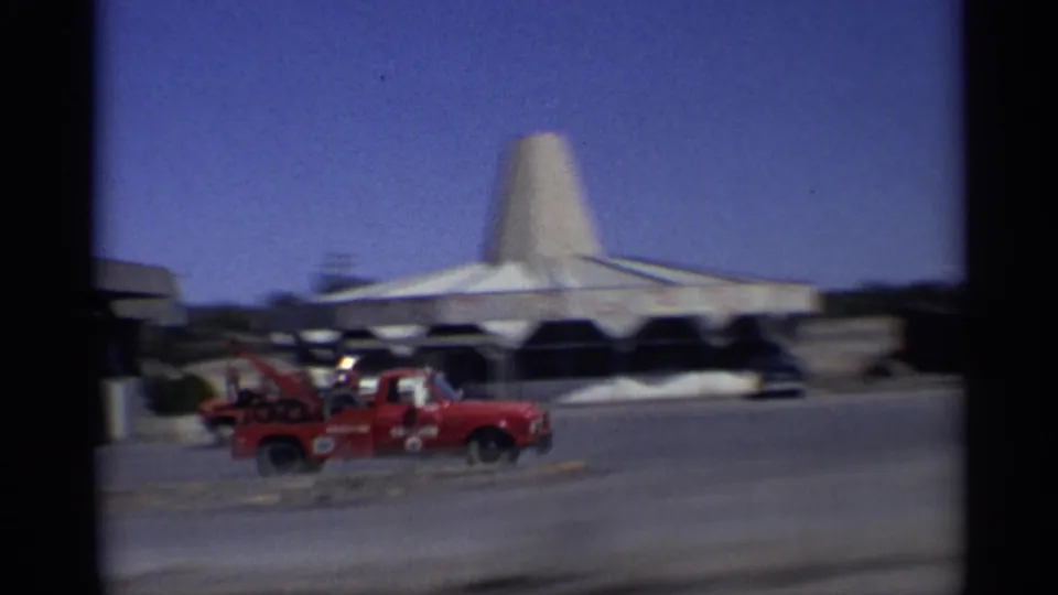 1959 Arizona home-movie frame showing a red tow truck, parked cars, and a distinctive conical-roof building beside a road.