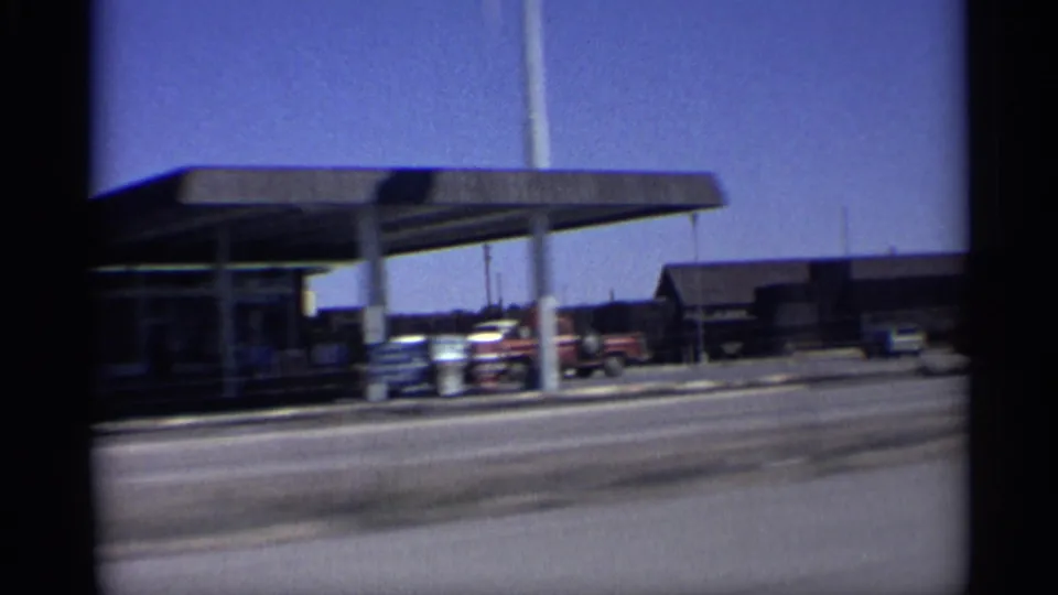 Arizona road-trip frame with a roadside canopy, parked vehicles, low buildings, and open sky seen from a moving car.