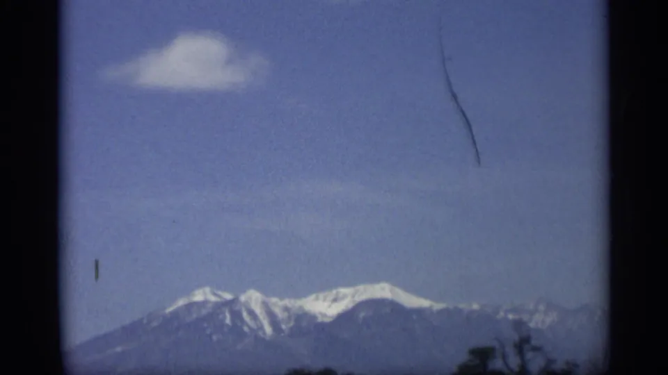 Zoomed 1959 Arizona home-movie view of a snow-capped mountain range, blue sky, sparse trees, and visible film edge marks.