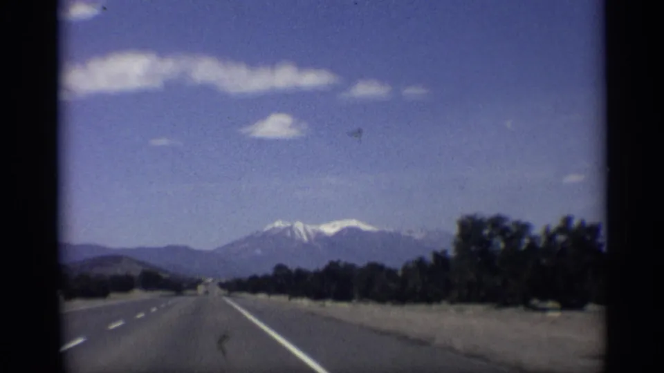 1959 Arizona 8mm road-view frame showing a highway receding toward snow-capped mountains with trees along the right side.