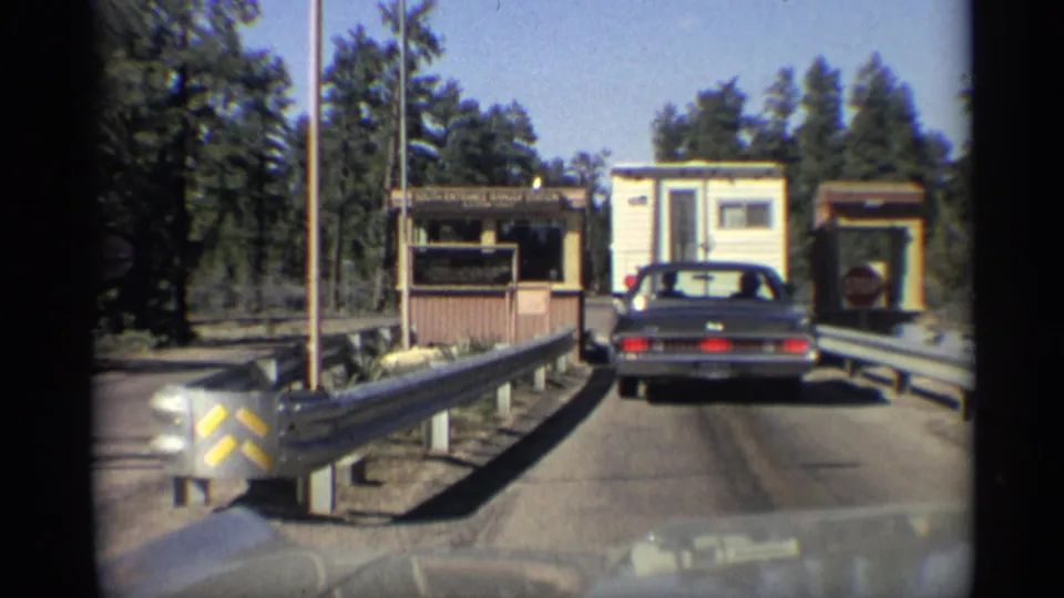 1959 Arizona road-trip frame showing a dark car at a small toll or checkpoint booth, with guardrails and trees nearby.