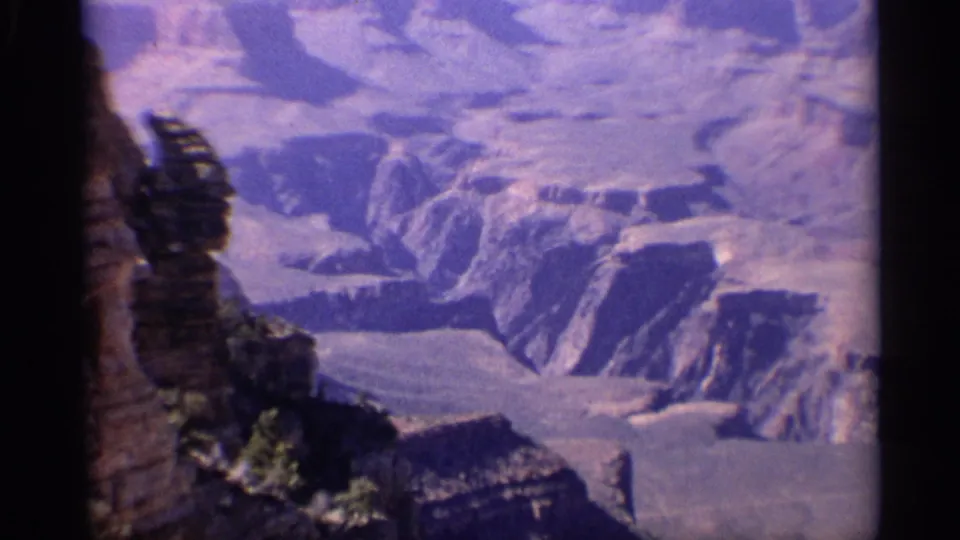 1959 8mm aerial view over the Grand Canyon in Arizona, showing layered cliffs, ravines, and soft purple-blue color shift.