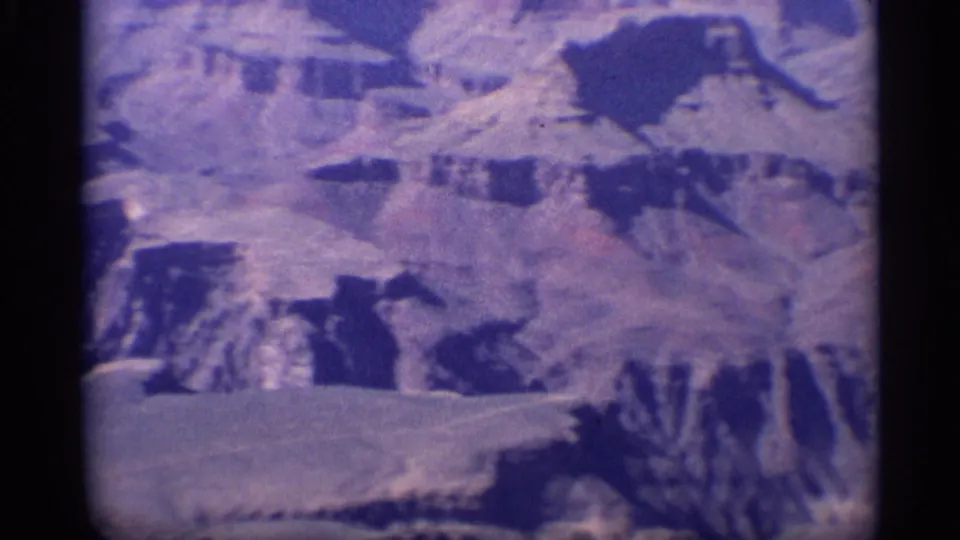 1959 8mm frame of the Grand Canyon from above, with pale plateau ledges, dark ravines, and rounded home-movie frame corners.