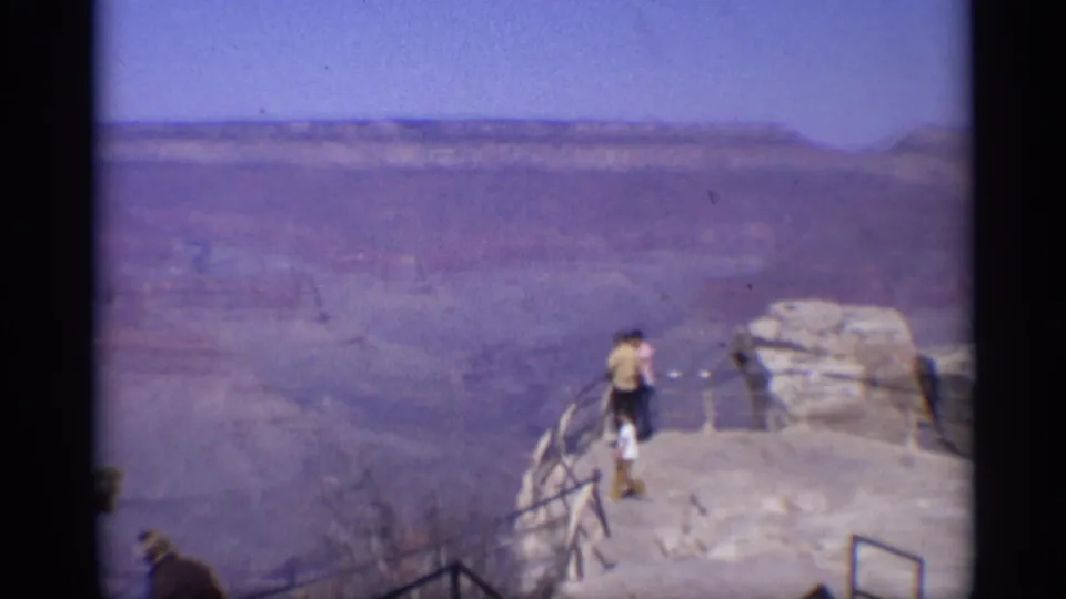1959 8mm frame showing two visitors on a rocky Grand Canyon viewpoint with railings and a wide canyon backdrop.