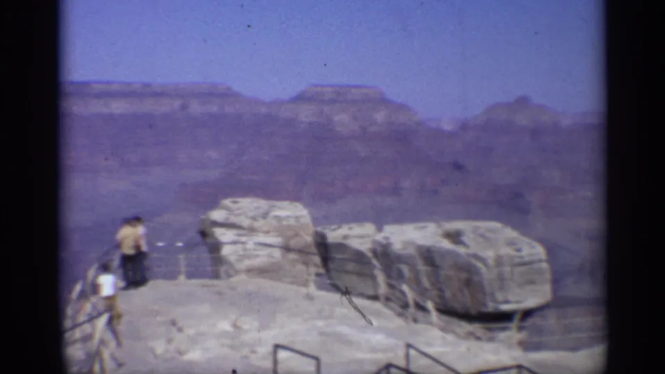 Vintage 8mm Arizona travel frame of visitors beside an overlook railing, with large rocks and Grand Canyon walls in view.