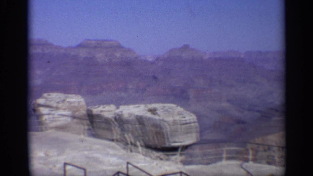 Canyon overlook with pale rock ledges, railing, distant mesas, and blue sky.
