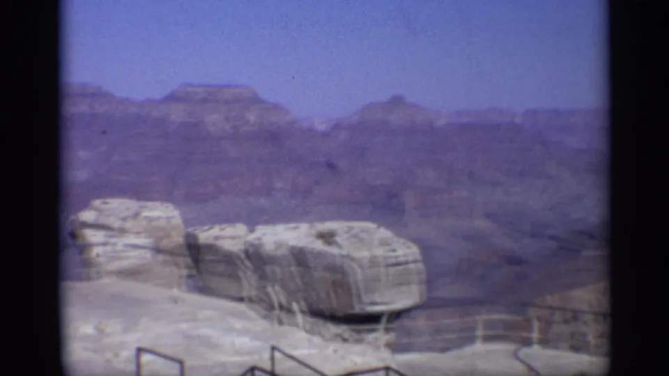 1959 Arizona 8mm frame showing a canyon overlook with pale rock ledges, railing, distant layered mesas, and blue sky.