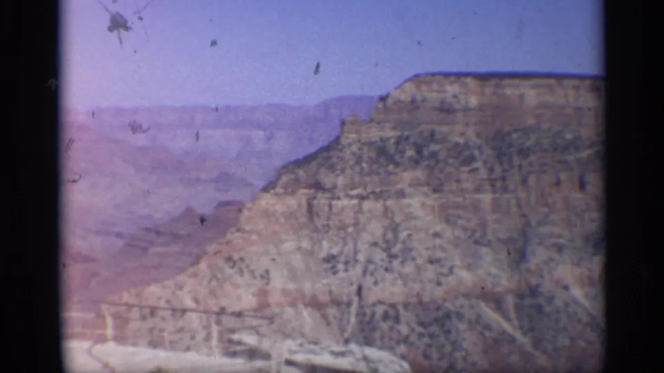 1959 Arizona 8mm frame showing a broad layered canyon cliff with distant haze, blue sky, and visible film dust.