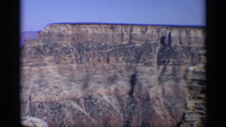 1959 Arizona home-movie frame showing distant layered canyon cliffs and rocky crevices beneath a pale blue sky.