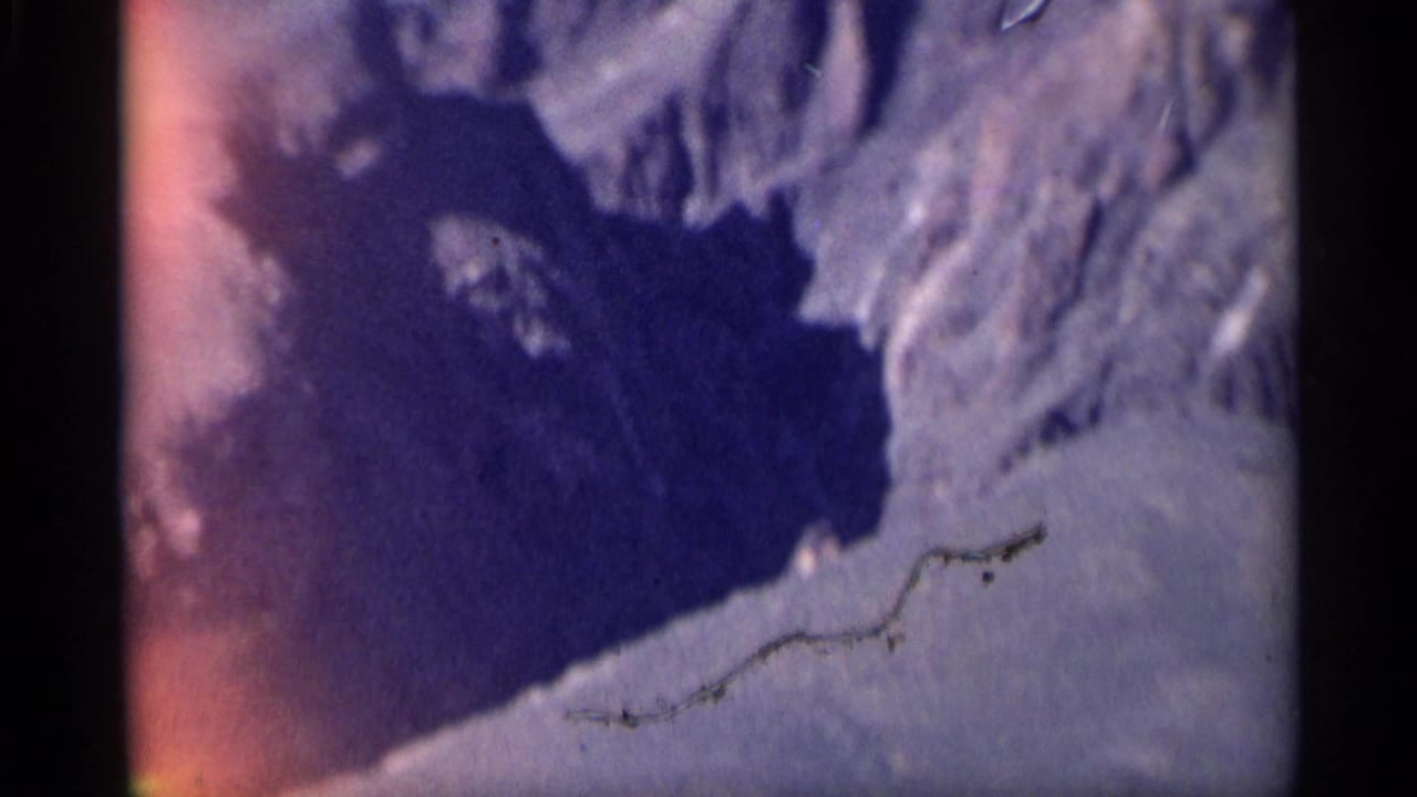 1959 Arizona 8mm frame showing a shadowed canyon basin bordered by dry pale rock and rugged desert terrain.