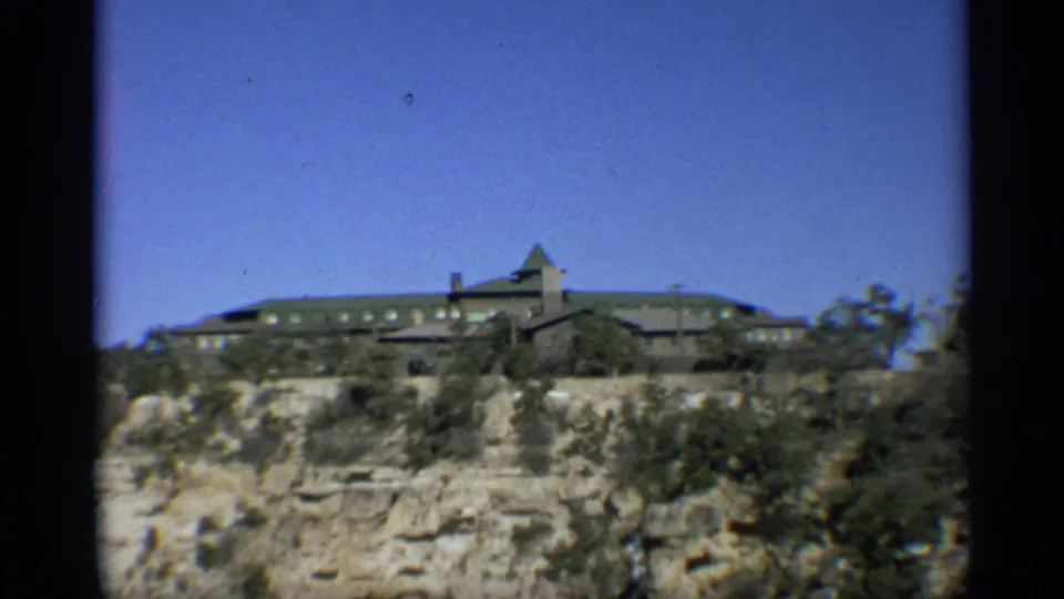 Large lodge-like structure on the Grand Canyon rim in Arizona, with scrub vegetation and layered rock below in a 1959 8mm frame.
