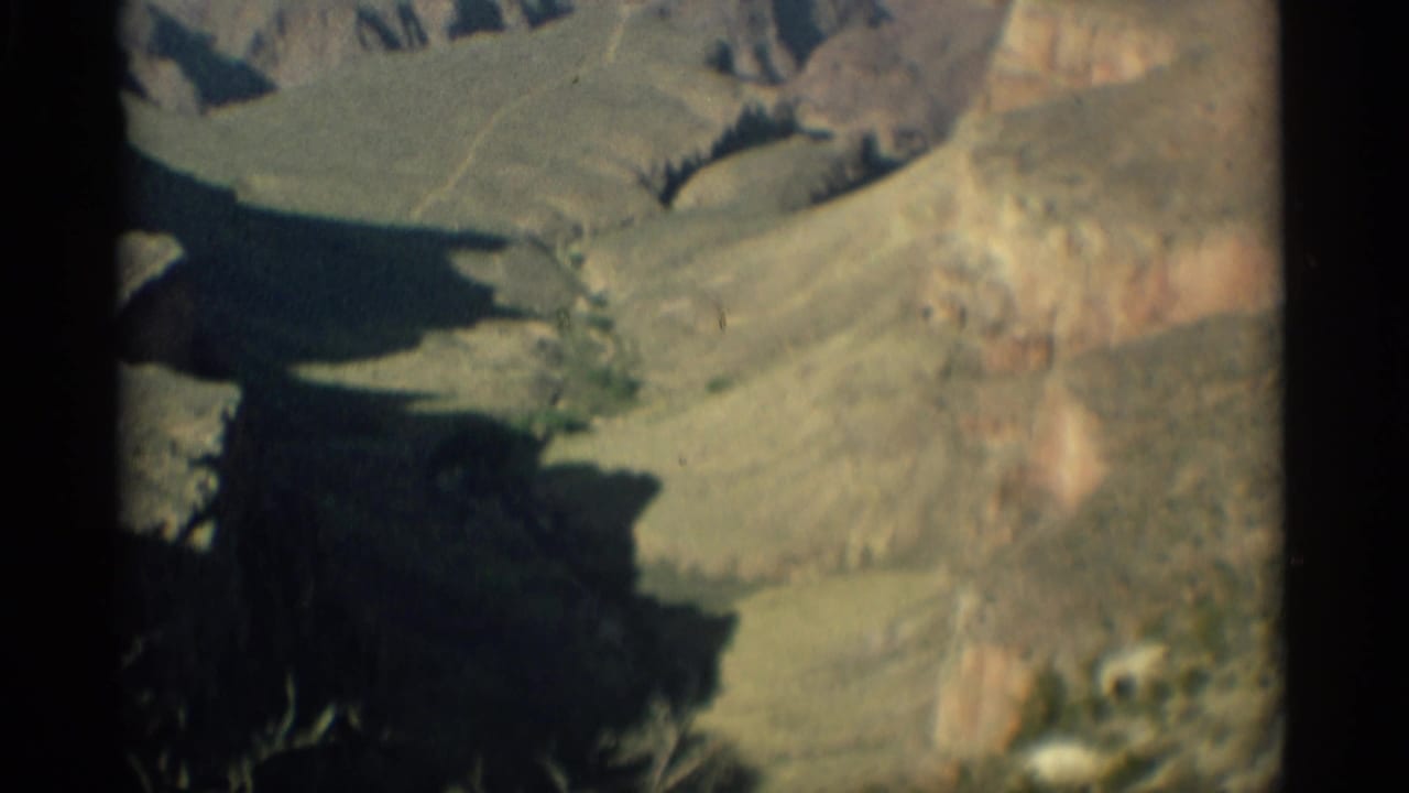 1959 Arizona aerial frame looking over canyon slopes, shadowed cuts, pale rock ridges, and muted green desert vegetation.