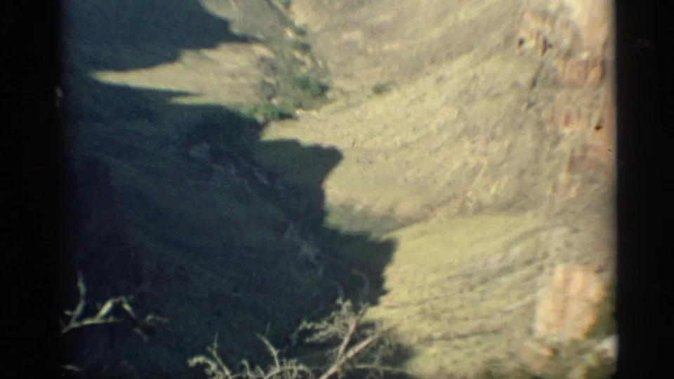 1959 Arizona 8mm frame showing a shadowed canyon edge, pale desert slopes, and scattered brush from an aerial viewpoint.