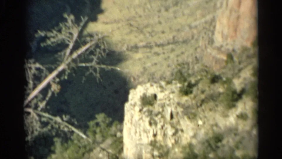 1959 Arizona aerial frame with rocky canyon ground, sparse shrubs, pale stone, and a strong shadow across the landscape.