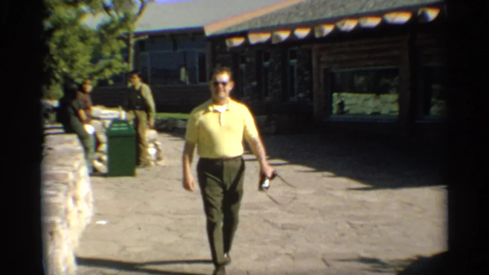 1959 Arizona home-movie frame of a man in sunglasses and a yellow shirt walking near a rustic log-style building.