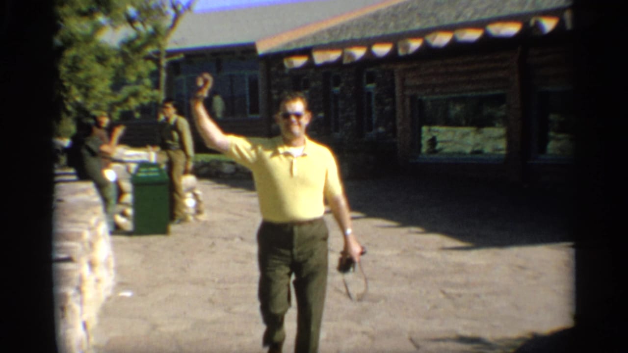 Man in yellow polo waves while walking near a rustic cabin and stone wall.