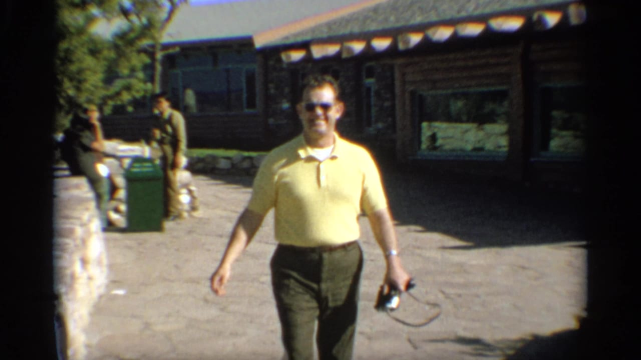 Man walks toward the camera holding a small camera near a cabin entrance.