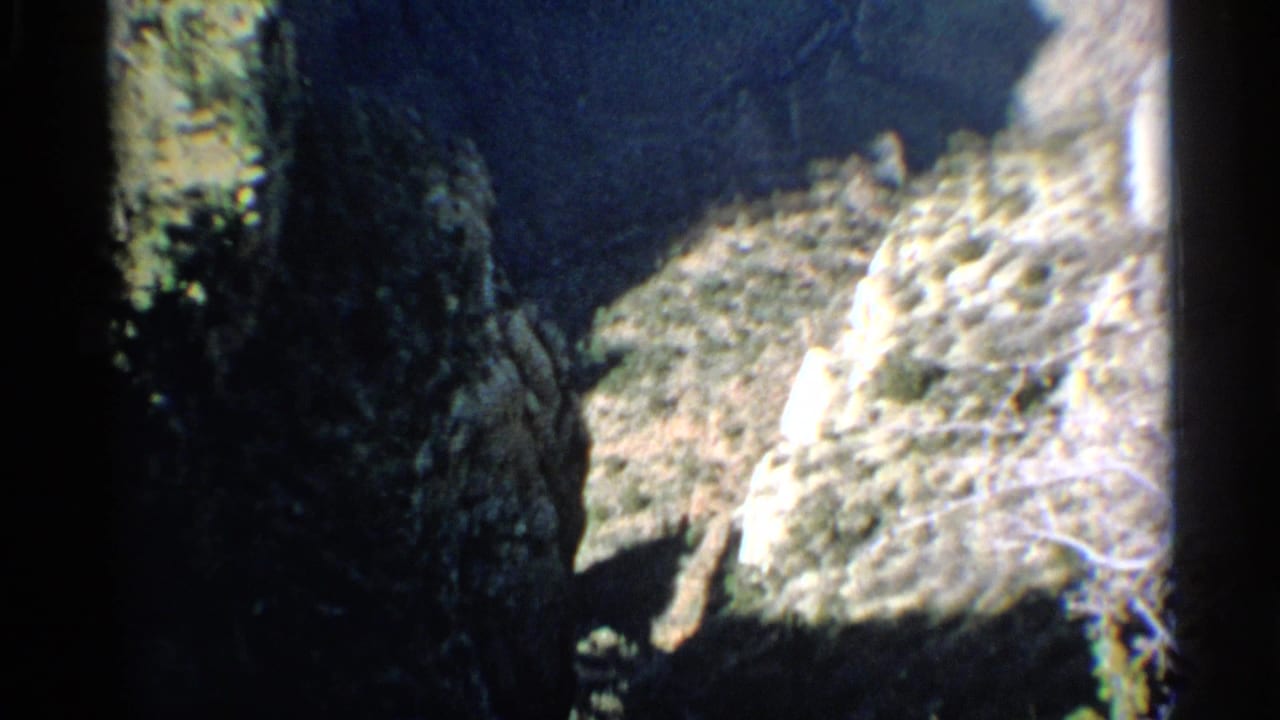 Dark rocky canyon wall with pale cliff face and sparse brush in soft 8mm color.