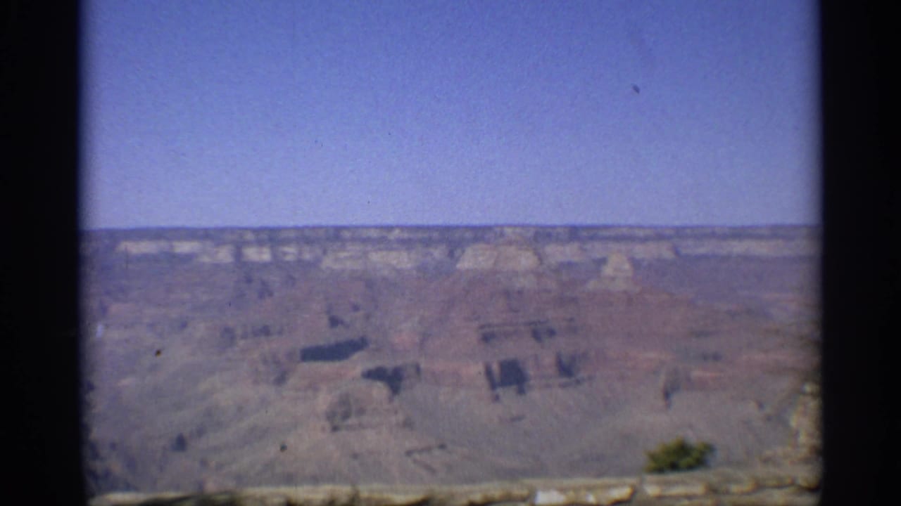 Distant canyon landscape with blue sky, pale rock formations, and dark vertical film edge.