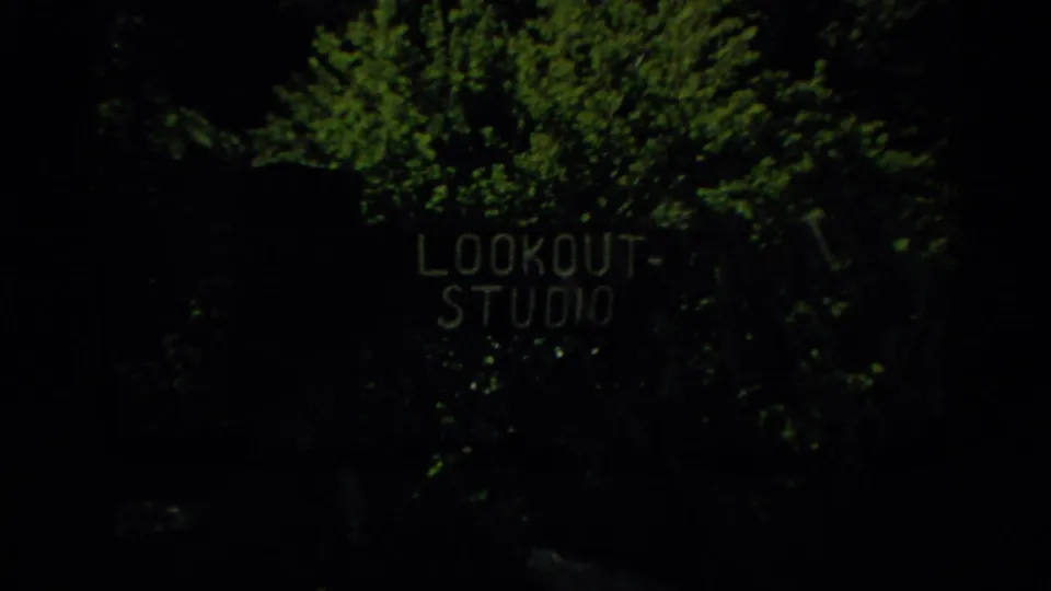 Dark 1959 8mm frame with green vegetation and faint sign-like lettering that appears to read LOOKOUT STUDIO.