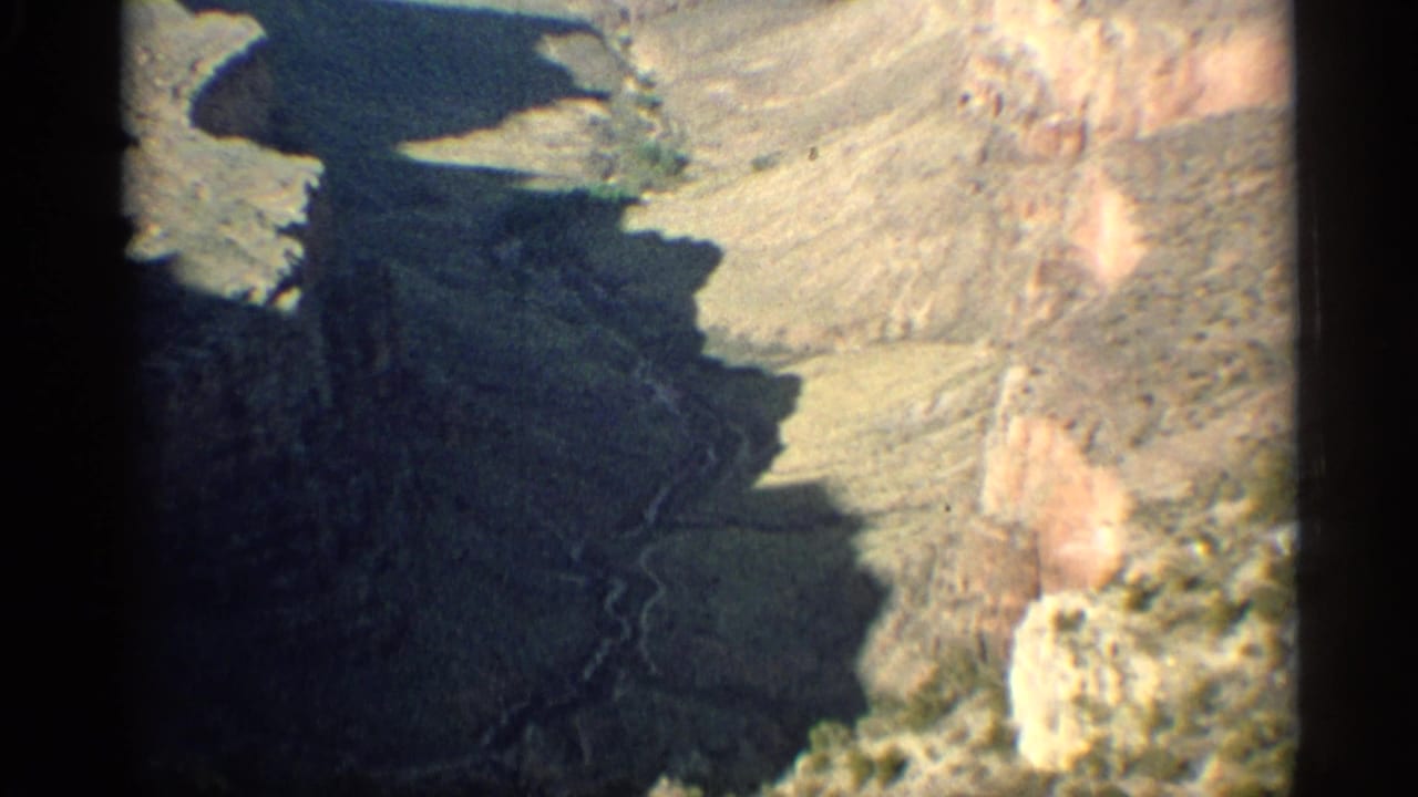 1959 Arizona 8mm aerial view looking down over dark canyon shadows, tan rock faces, and sparse desert terrain.