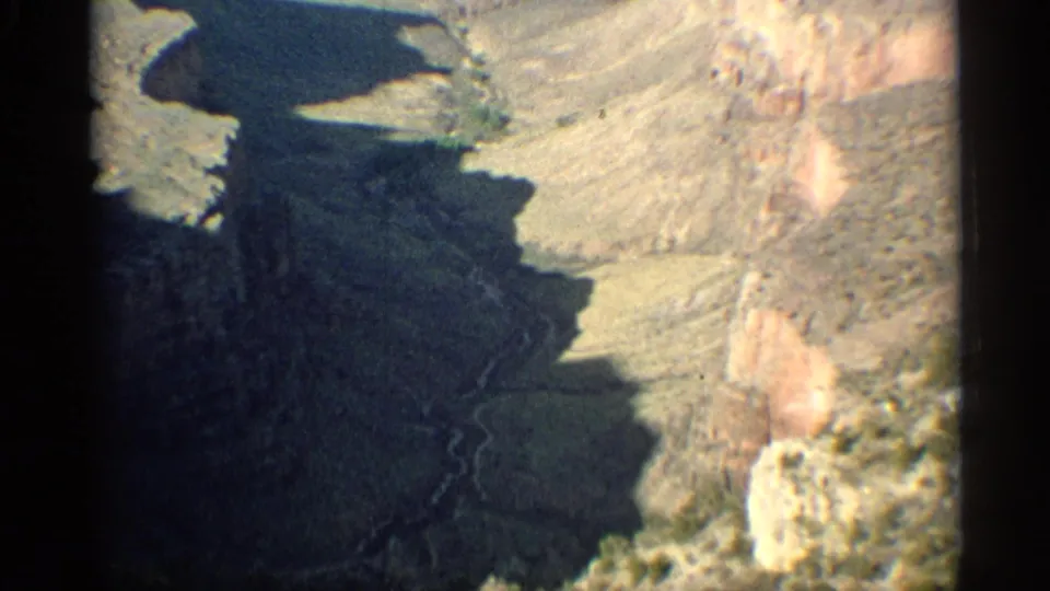 1959 Arizona 8mm aerial view looking down over dark canyon shadows, tan rock faces, and sparse desert terrain.