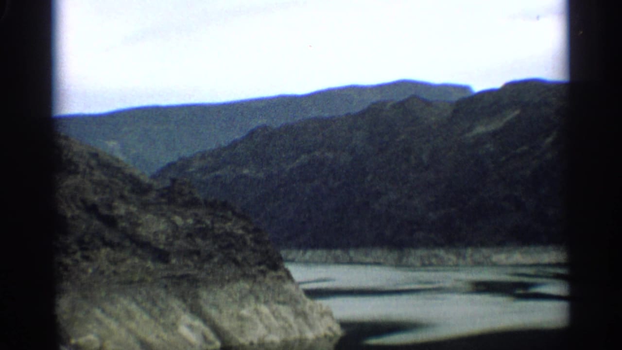 Rocky mountain slope above pale water with distant ridges under a bright sky.