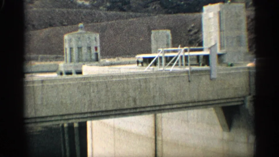 1959 8mm frame showing Hoover Dam deck details, metal railings, a small cylindrical structure, concrete wall, and canyon background.