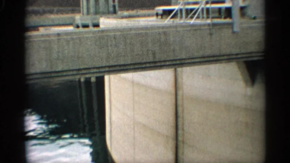Close 1959 8mm view of Hoover Dam concrete face and railings, with a tower section, reservoir water, and desert canyon behind.