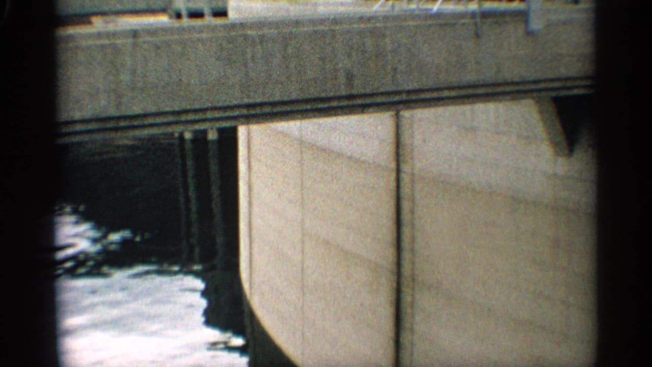 Concrete dam wall beside churning water, framed close from above.